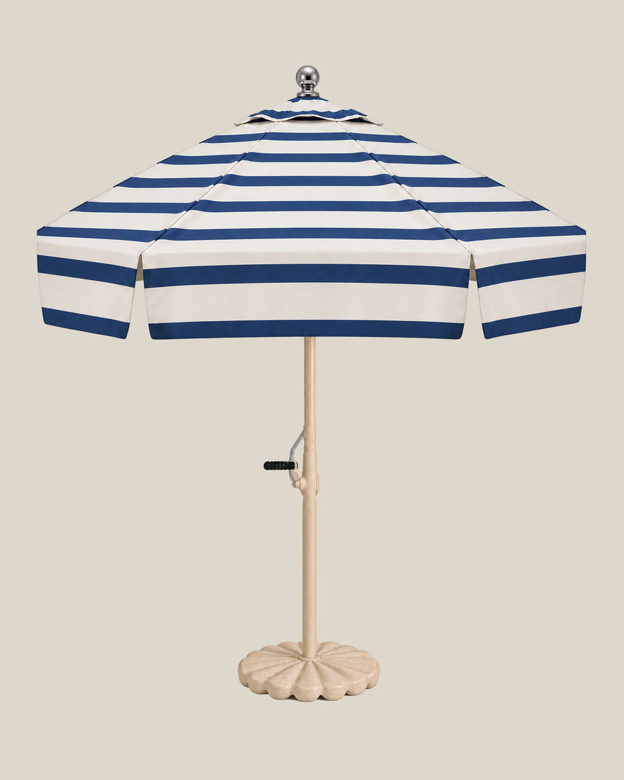 Riviera umbrella with navy stripes across its ivory canopy and tiered top, beige pole and scalloped travertine base on bone