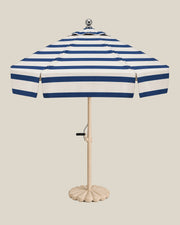 Riviera umbrella with navy stripes across its ivory canopy and tiered top, beige pole and scalloped travertine base on bone